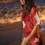 A woman wearing a red and white abstract print cover-up dress walking on a beach at sunset.
