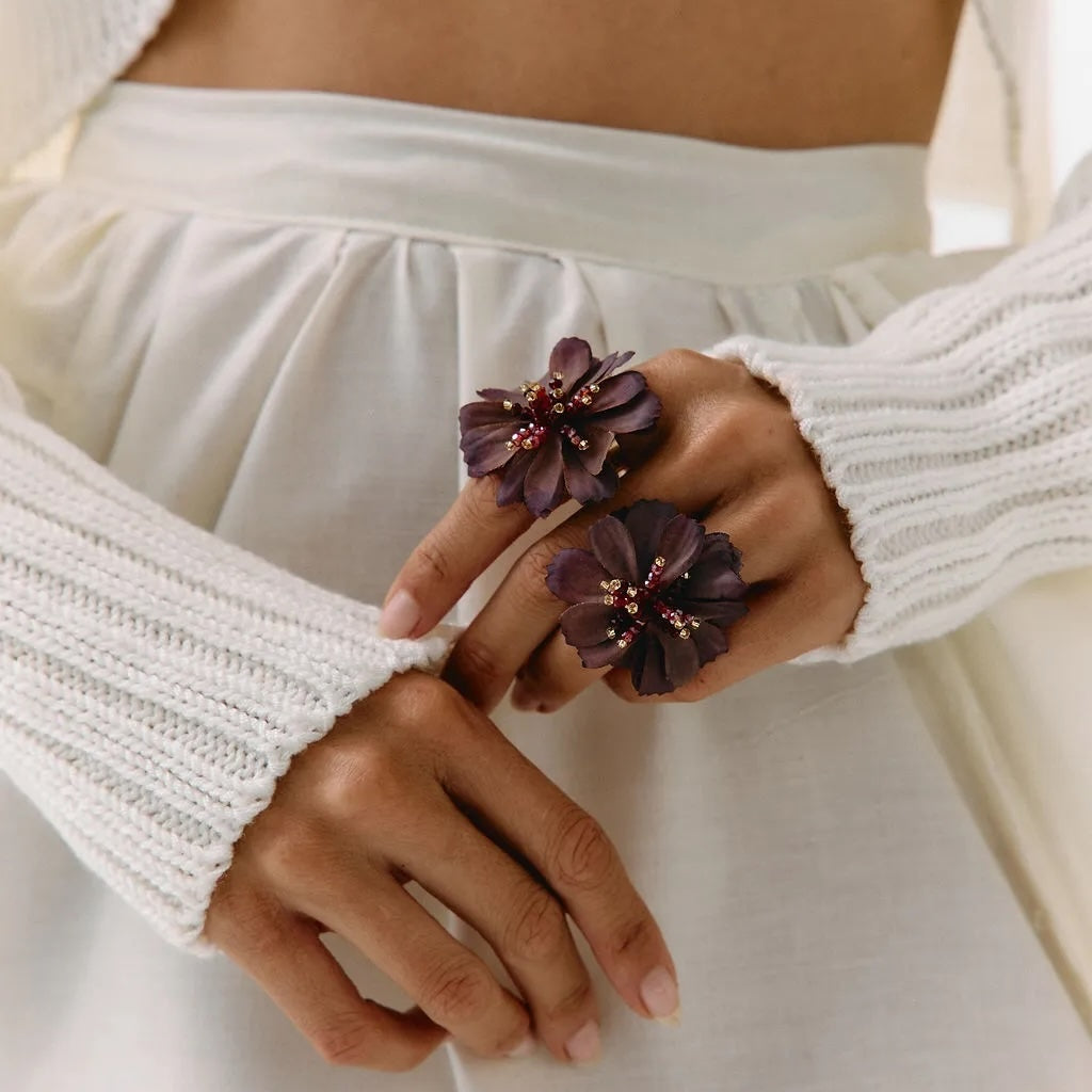 A close-up of a person wearing a white ribbed knit cropped sweater and white skirt, holding their hands together to display a pair of large dark purple flower-shaped rings with small pink bead accents.