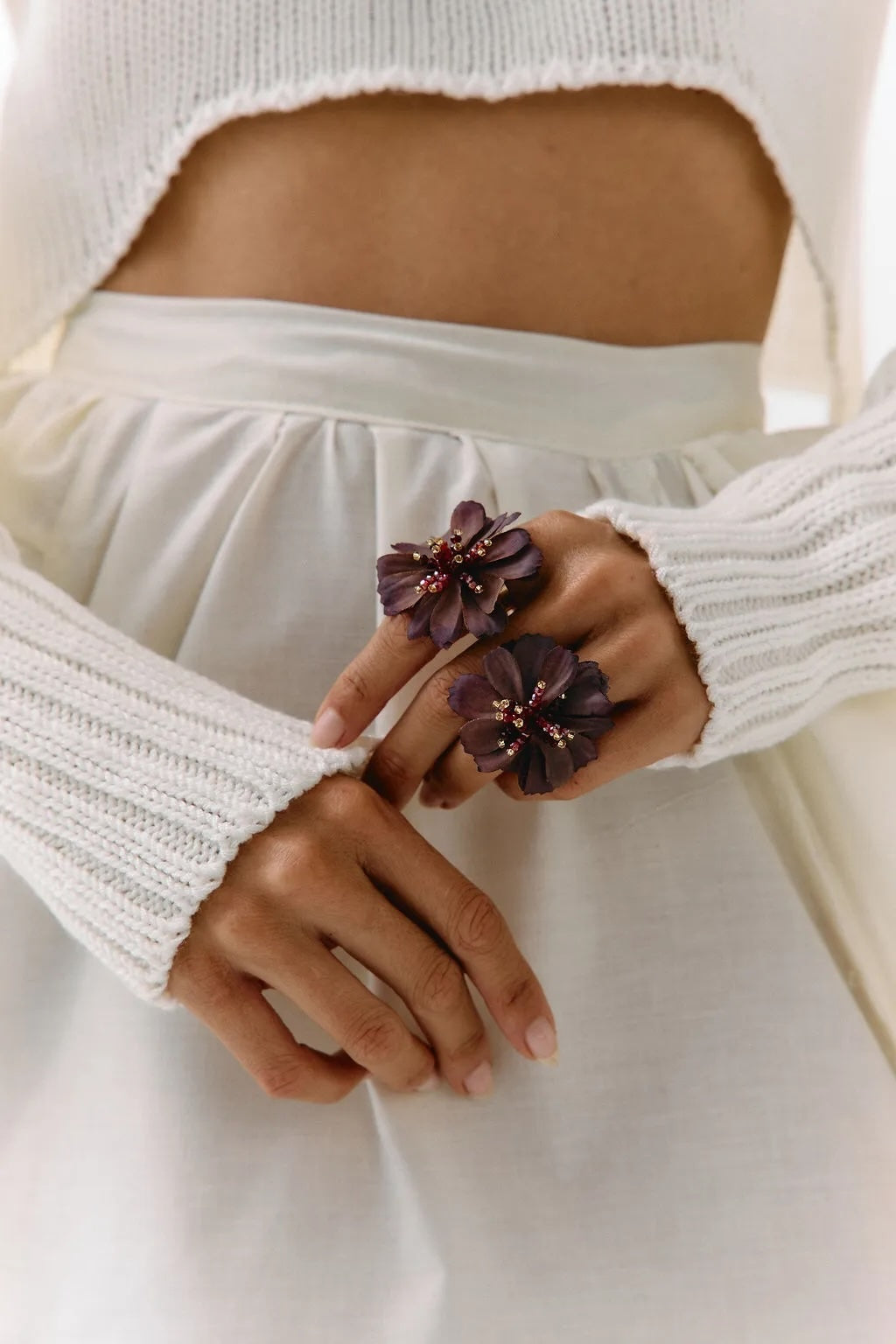 A close-up of a person wearing a white ribbed knit cropped sweater and white skirt, holding their hands together to display a pair of large dark purple flower-shaped rings with small pink bead accents.