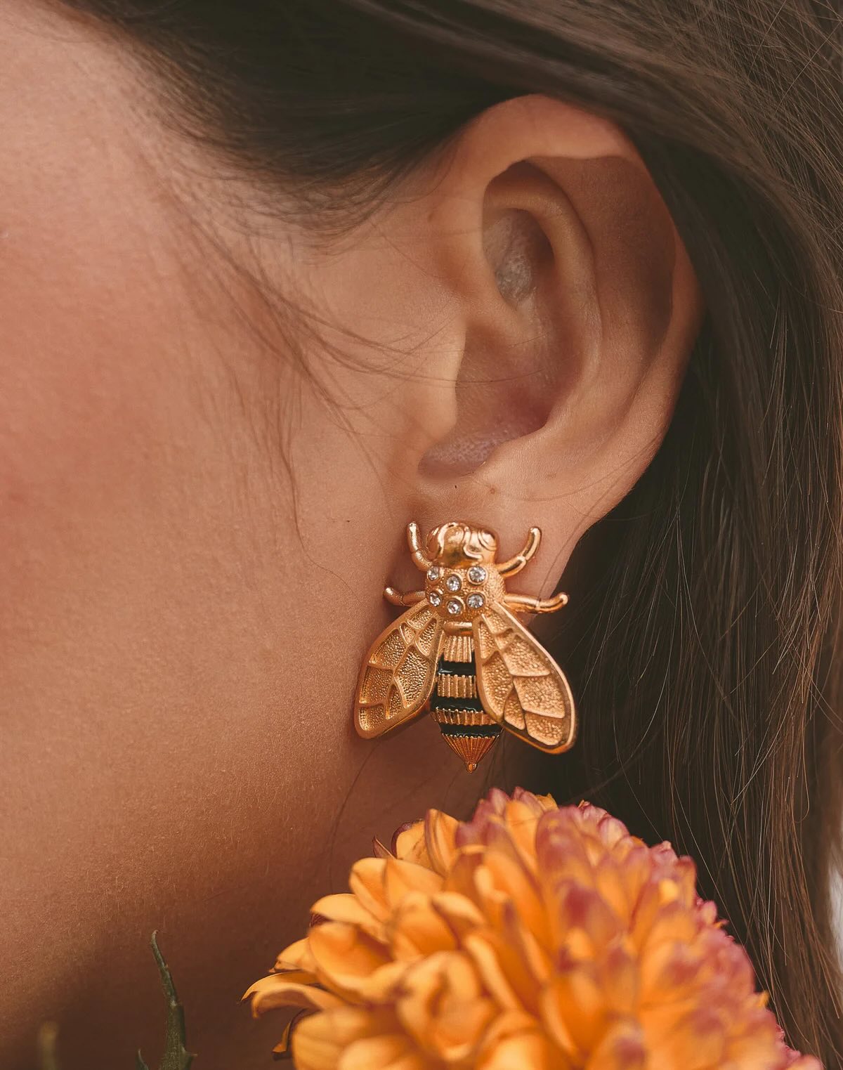 Close-up of an ear wearing a gold bee-shaped earring with an orange flower in the foreground.