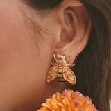 Close-up of an ear wearing a gold bee-shaped earring with an orange flower in the foreground.