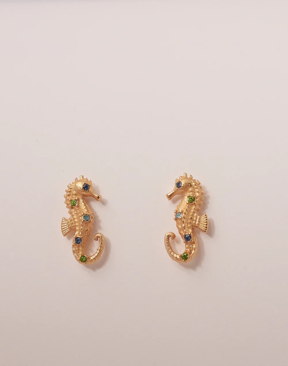 Gold seahorse earrings with green gemstones on a beige background