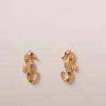 Gold seahorse earrings with green gemstones on a beige background