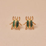 Gold beetle-shaped earrings with green accents on a beige background
