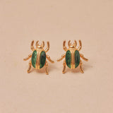 Gold beetle-shaped earrings with green accents on a beige background