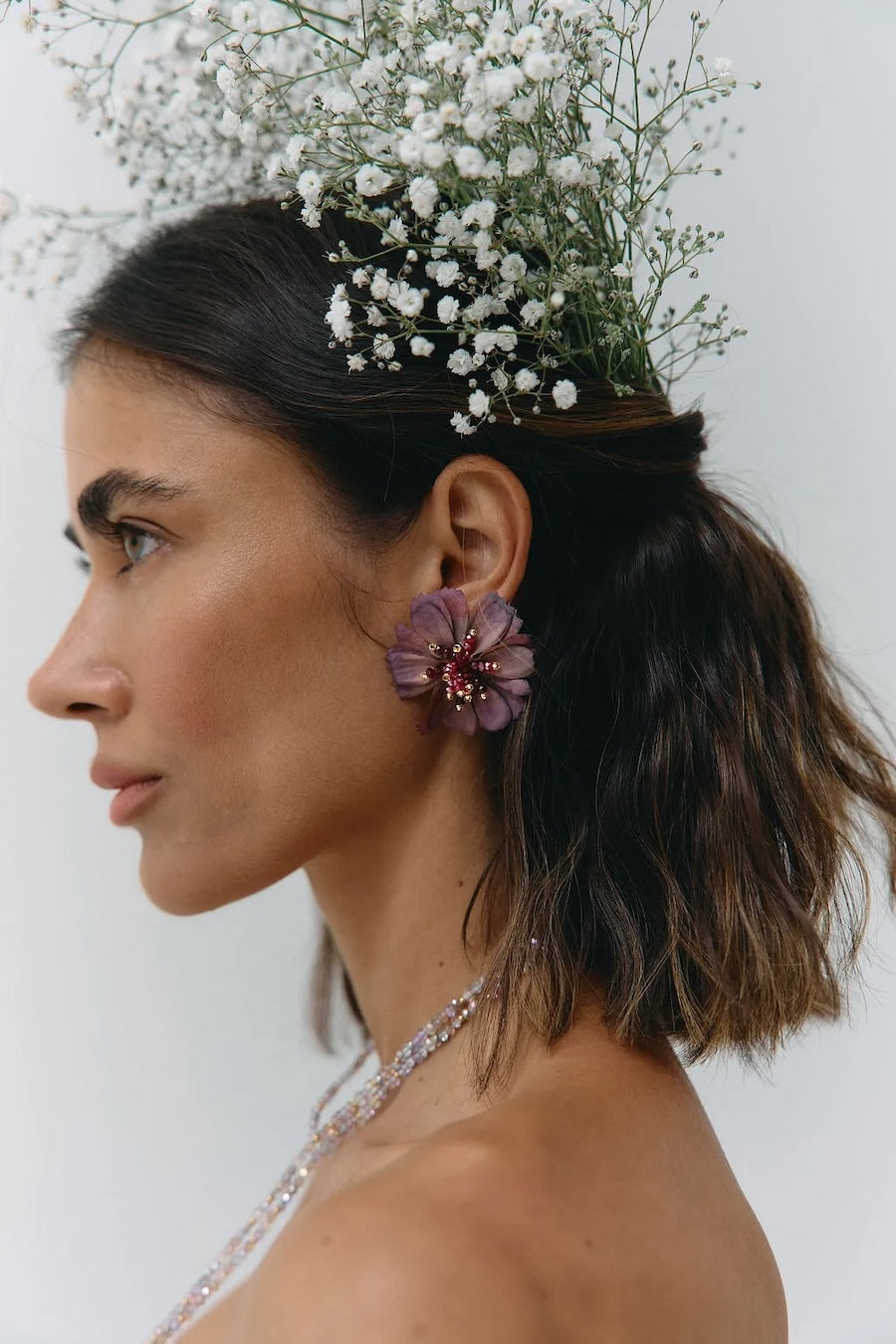 A woman in profile wearing a floral headpiece made of small white baby's breath and a purple flower earring with a red center, against a plain light background.