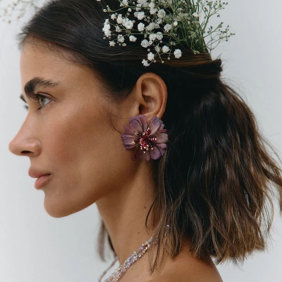 A woman in profile wearing a floral headpiece made of small white baby's breath and a purple flower earring with a red center, against a plain light background.