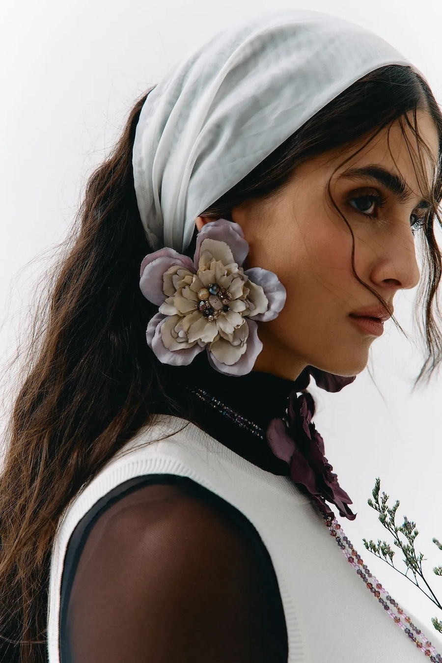 A close-up portrait of a woman wearing a white headscarf, a large lavender and cream fabric flower accessory attached near her ear, and a white sleeveless top against a plain light background.