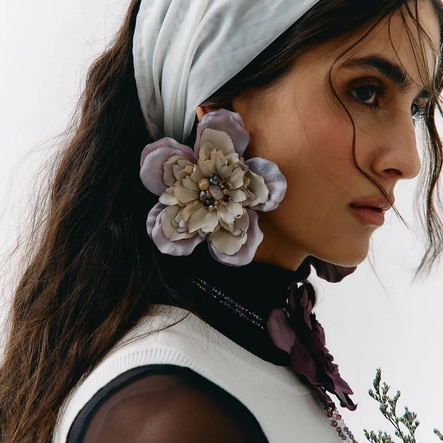 A close-up portrait of a woman wearing a white headscarf, a large lavender and cream fabric flower accessory attached near her ear, and a white sleeveless top against a plain light background.