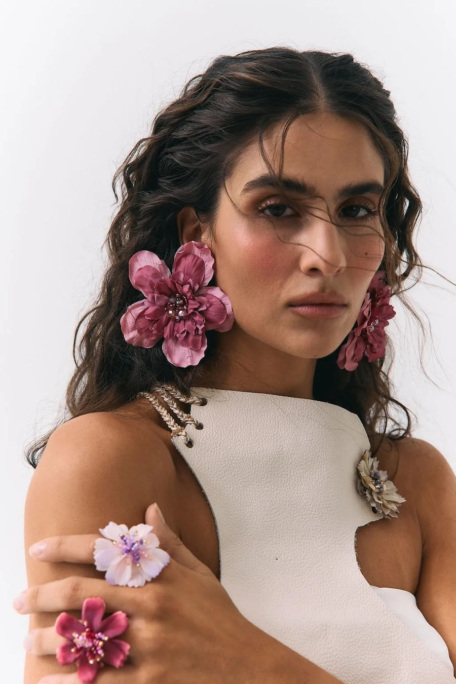 A close-up portrait of a woman wearing large pink flower-shaped earrings, matching flower rings on her fingers, and a white sleeveless top with floral embellishments against a plain light background.