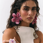 A close-up portrait of a woman wearing large pink flower-shaped earrings, matching flower rings on her fingers, and a white sleeveless top with floral embellishments against a plain light background.