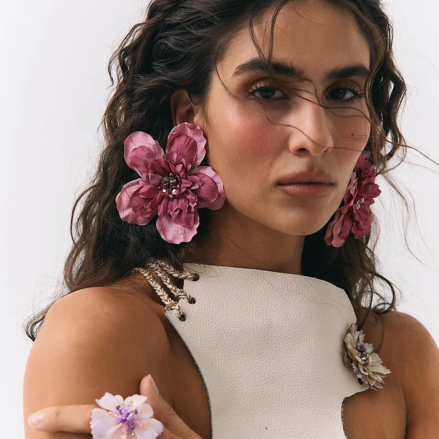 A close-up portrait of a woman wearing large pink flower-shaped earrings, matching flower rings on her fingers, and a white sleeveless top with floral embellishments against a plain light background.
