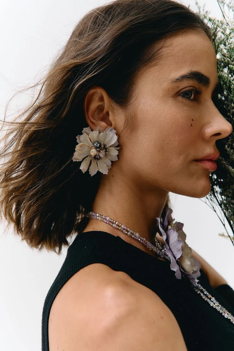 A close-up side profile of a woman with short brown hair wearing a large cream flower-shaped earring and a matching floral necklace with a black sleeveless top.