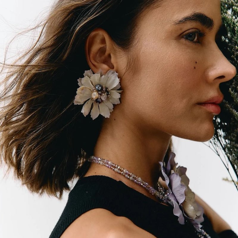 A close-up side profile of a woman with short brown hair wearing a large cream flower-shaped earring and a matching floral necklace with a black sleeveless top.