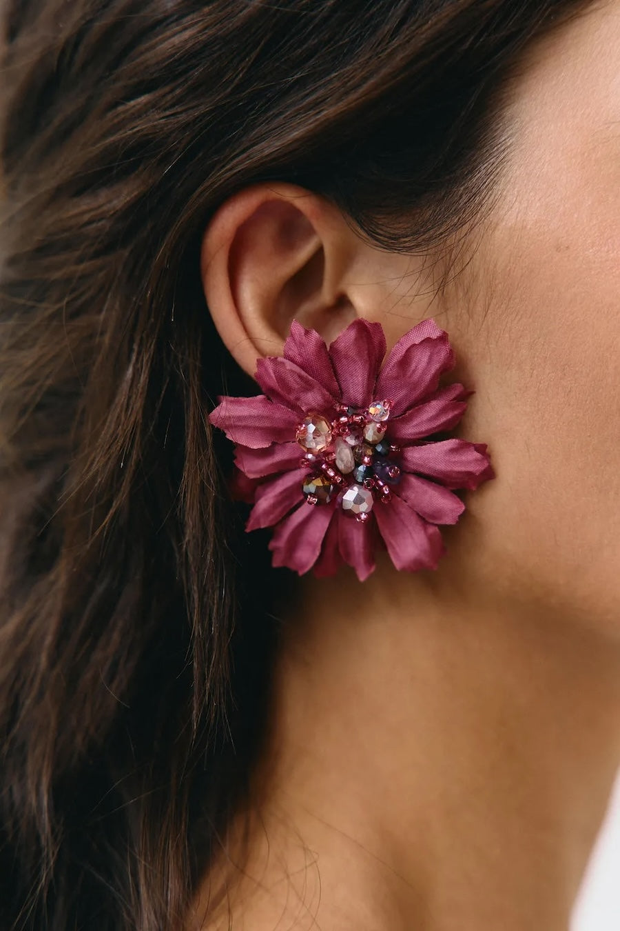 A close-up of a person's ear wearing a large, dark magenta flower-shaped stud earring adorned with colorful rhinestones in the center.