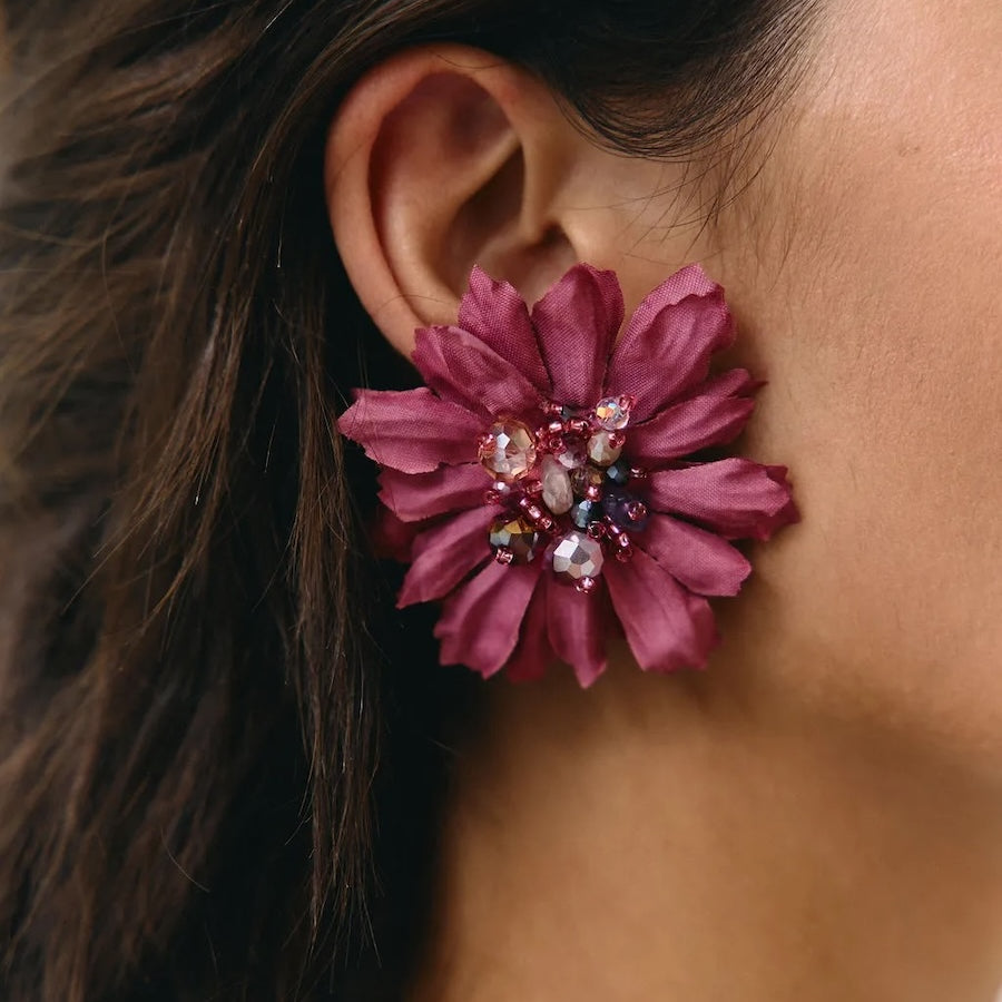 A close-up of a person's ear wearing a large, dark magenta flower-shaped stud earring adorned with colorful rhinestones in the center.
