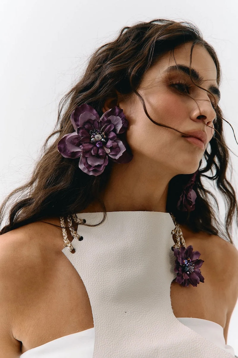 A close-up of a woman wearing large purple fabric flower drop earrings and matching flower-shaped earrings on her earlobes, paired with a white one-shoulder top against a plain light background.