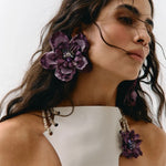 A close-up of a woman wearing large purple fabric flower drop earrings and matching flower-shaped earrings on her earlobes, paired with a white one-shoulder top against a plain light background.