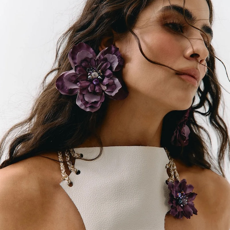 A close-up of a woman wearing large purple fabric flower drop earrings and matching flower-shaped earrings on her earlobes, paired with a white one-shoulder top against a plain light background.