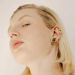 Woman wearing gold earrings with green gemstones on a plain background.