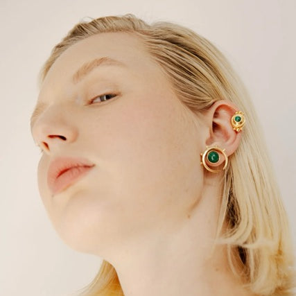 Woman wearing gold earrings with green gemstones on a plain background.