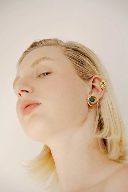 Woman wearing gold earrings with green gemstones on a plain background.