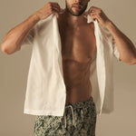 Man wearing patterned swim shorts and a white shirt over his shoulders against a beige background.
