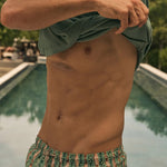 Man in green shirt and fish patterned shorts standing by a pool with trees in the background.