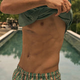 Man in green shirt and fish patterned shorts standing by a pool with trees in the background.