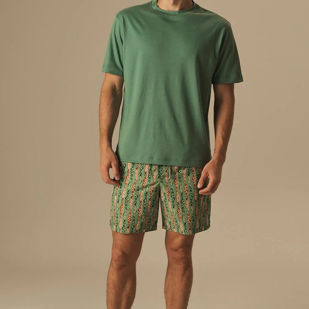 Man wearing a green t-shirt and fish patterned swim shorts on a beige background.