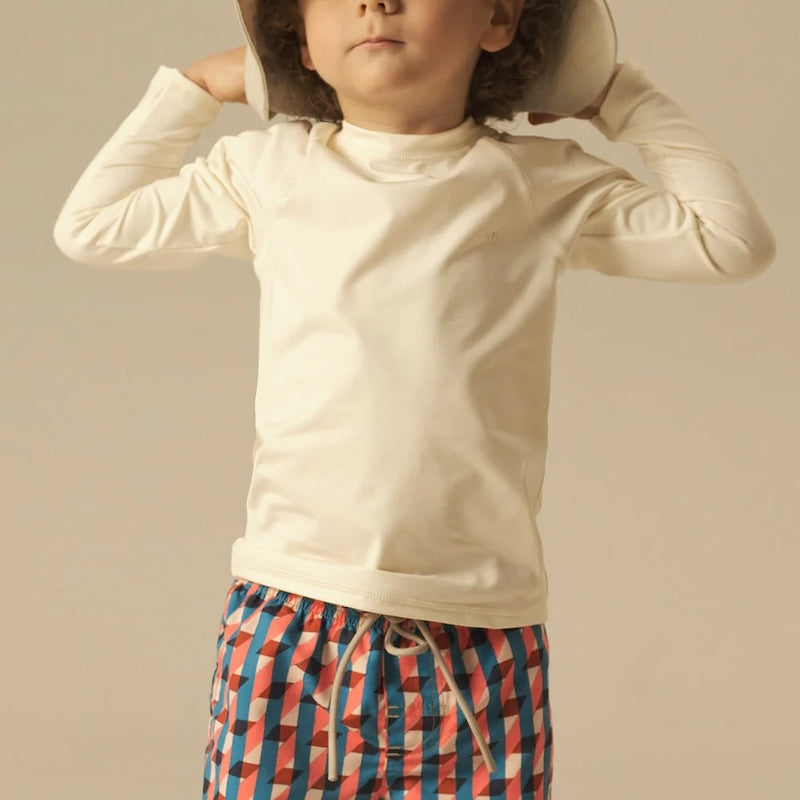 Child wearing a beige long-sleeve shirt and colorful geometric swim shorts against a neutral background.