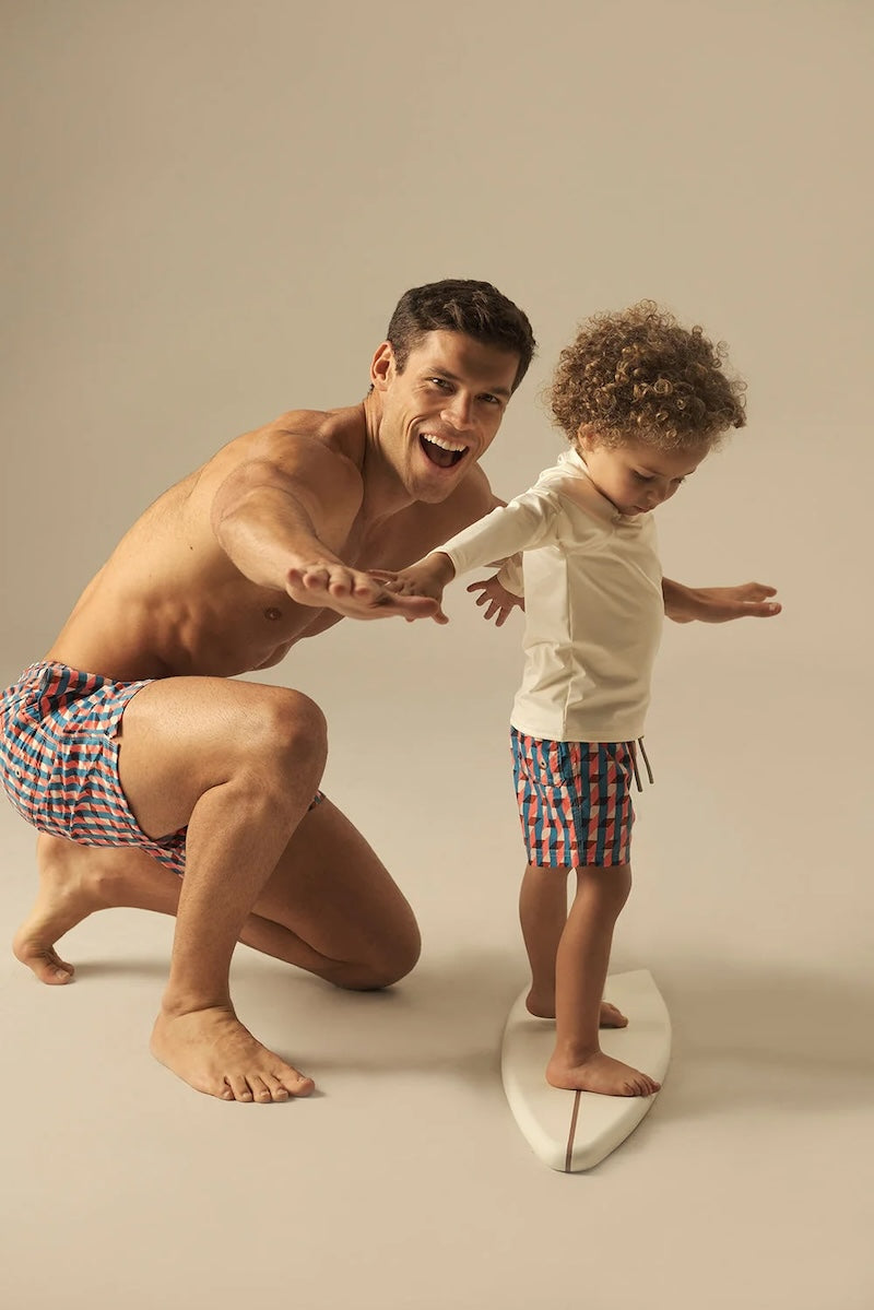Man and child in matching geometric swim shorts with surfboard against a plain background.