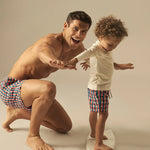 Man and child in matching geometric swim shorts with surfboard against a plain background.