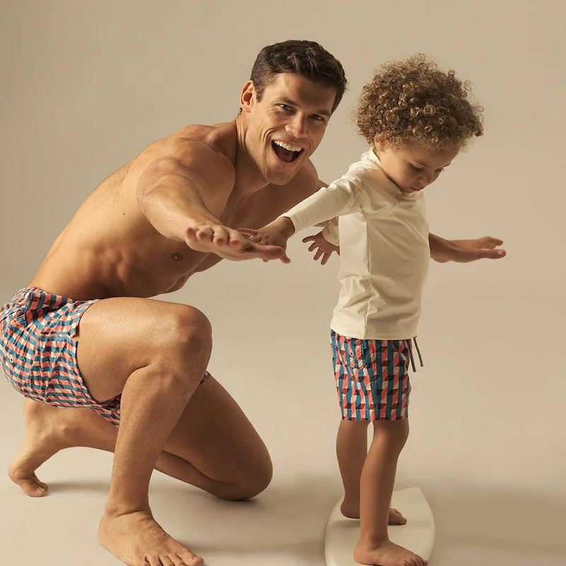 Man and child in matching geometric swim shorts with surfboard against a plain background.