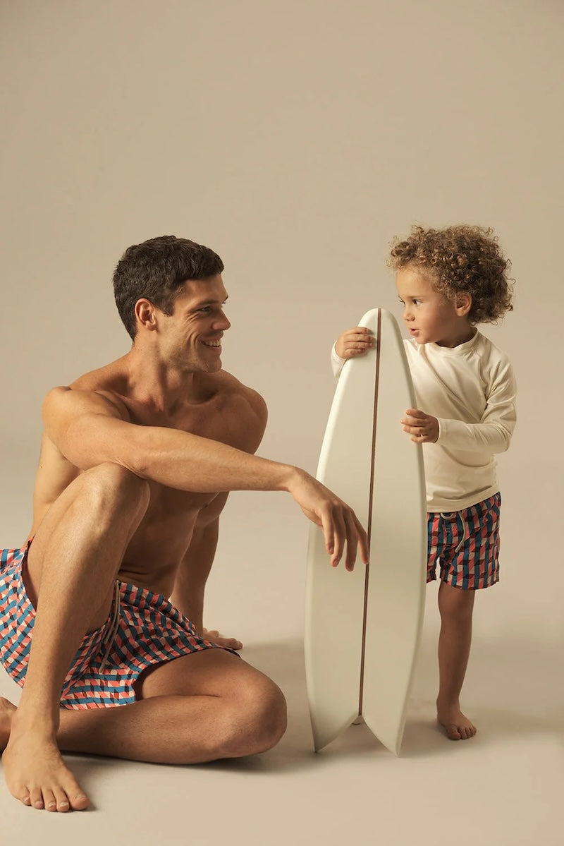 Man and child with a surfboard on a beige background wearing matching geometric swim shorts. 