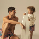 Man and child with a surfboard on a beige background wearing matching geometric swim shorts. 