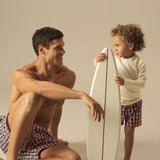 Man and child with a surfboard on a beige background wearing matching geometric swim shorts. 