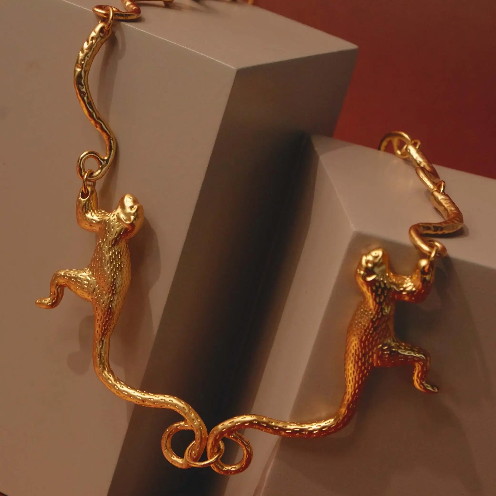 A 24K gold-plated necklace with a sculptural design featuring two Titi monkeys linked by their tails against a red background.