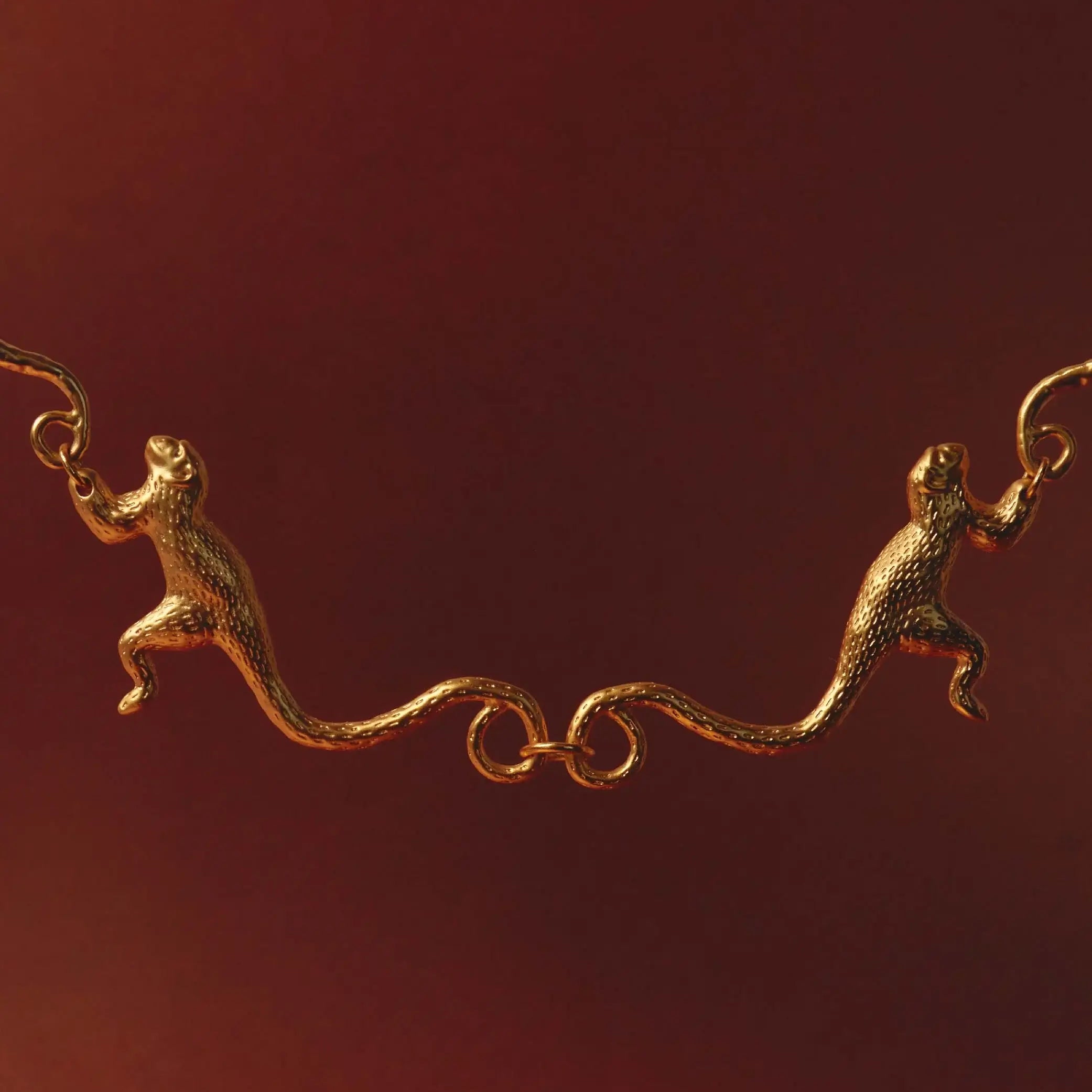 A 24K gold-plated necklace with a sculptural design featuring two Titi monkeys linked by their tails against a red background.