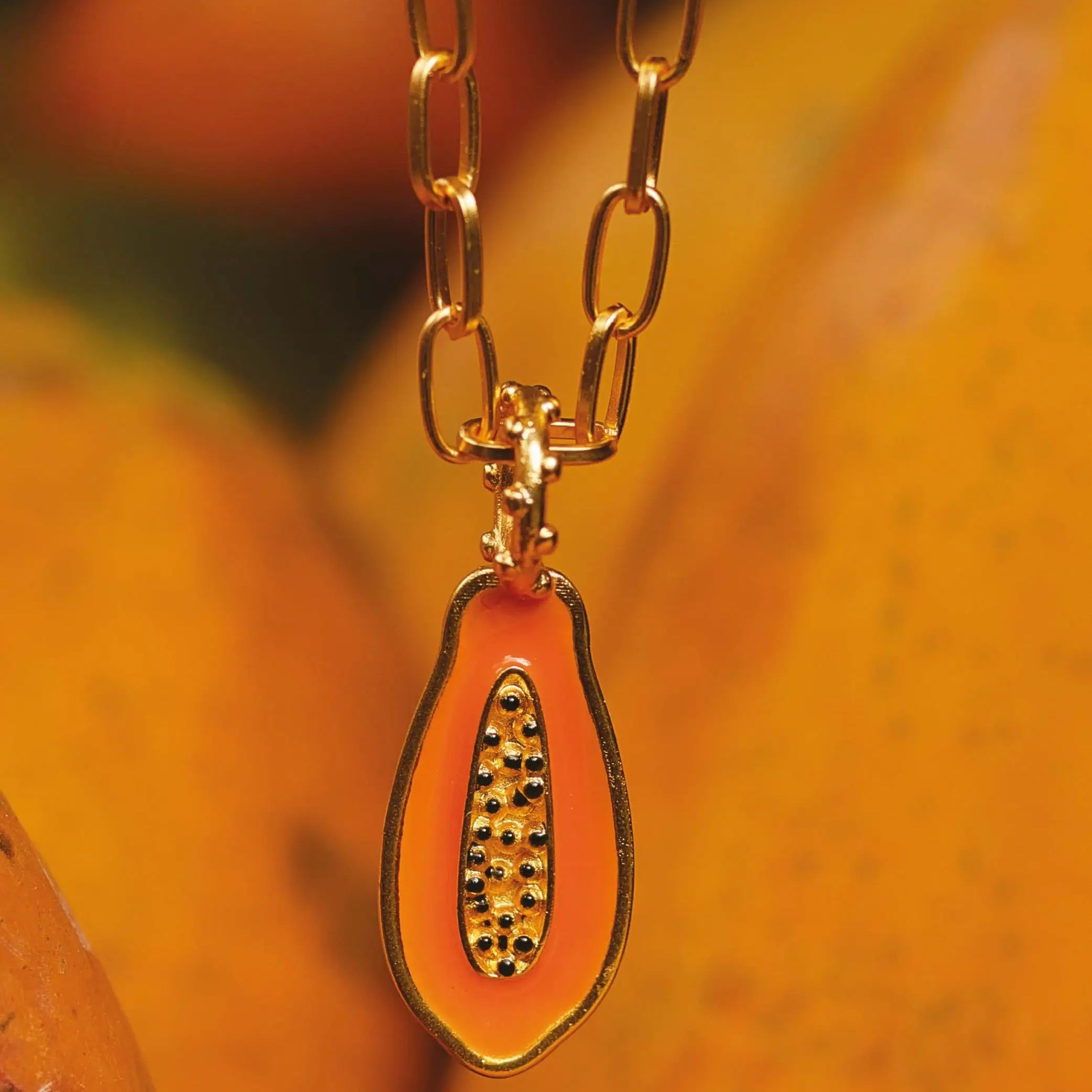 A gold-plated necklace with a papaya charm, displayed against an orange and yellow background.