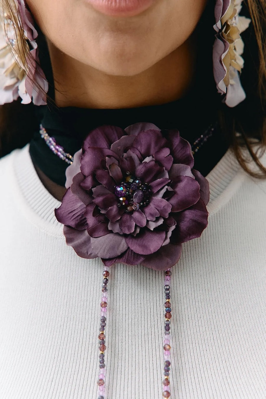 A close-up of a person wearing a large purple fabric flower brooch with dangling beaded chains over a white ribbed sweater.