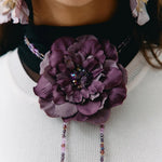 A close-up of a person wearing a large purple fabric flower brooch with dangling beaded chains over a white ribbed sweater.
