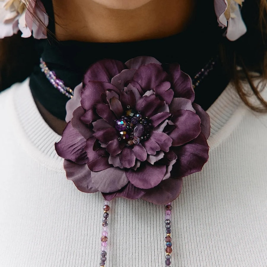 A close-up of a person wearing a large purple fabric flower brooch with dangling beaded chains over a white ribbed sweater.