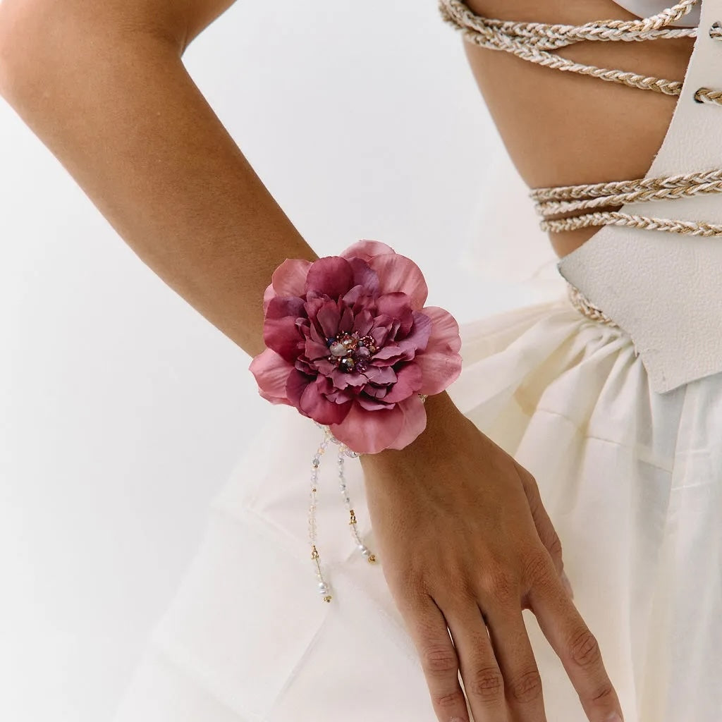 A close-up of a model wearing a large, dark pink peony flower wristband with delicate silver beaded chains draping down her forearm, paired with a white dress and gold chain corset details.