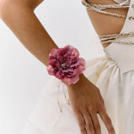 A close-up of a model wearing a large, dark pink peony flower wristband with delicate silver beaded chains draping down her forearm, paired with a white dress and gold chain corset details.
