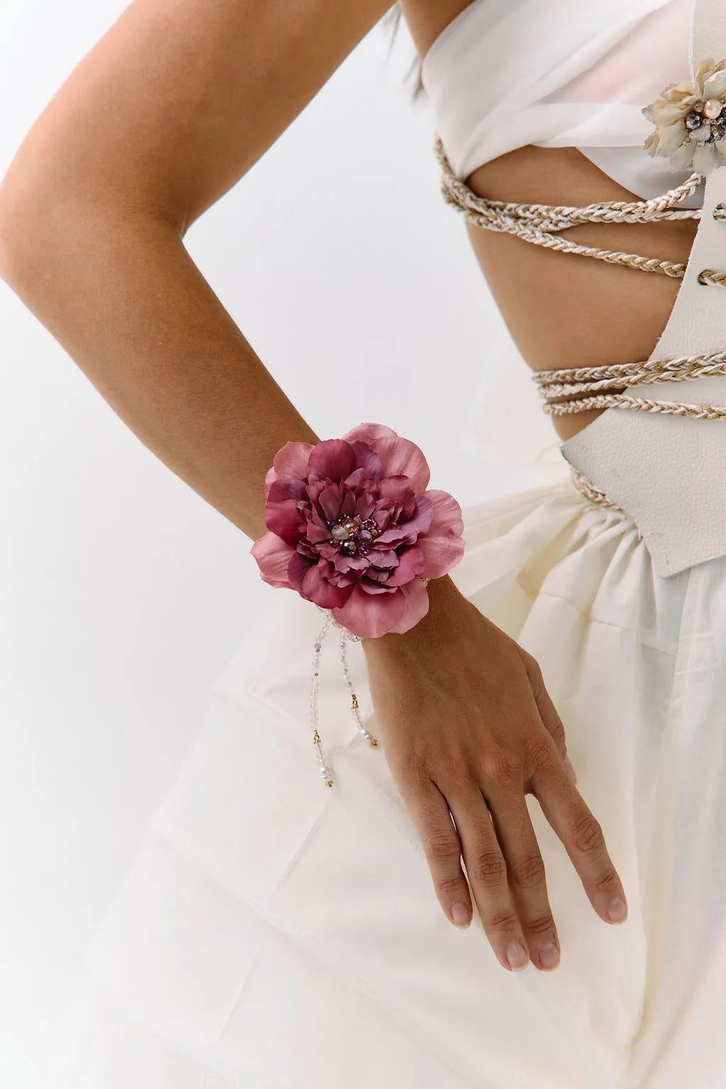A close-up of a model wearing a large, dark pink peony flower wristband with delicate silver beaded chains draping down her forearm, paired with a white dress and gold chain corset details.