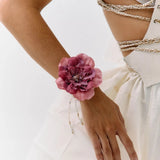 A close-up of a model wearing a large, dark pink peony flower wristband with delicate silver beaded chains draping down her forearm, paired with a white dress and gold chain corset details.
