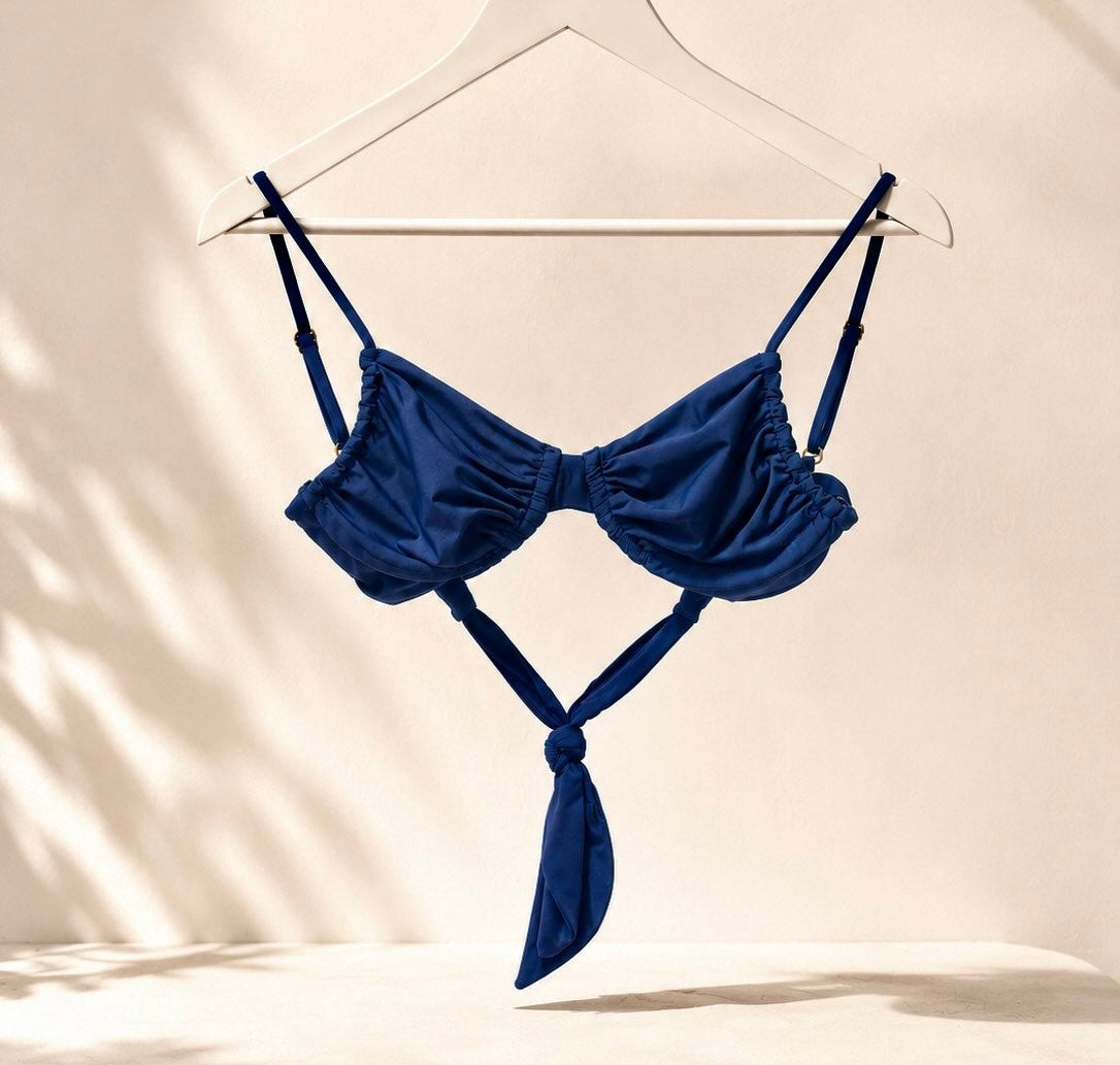 A navy blue strappy bikini top hanging on a white hanger from a gold clothing rack, set against a warm beige wall with soft leaf shadows.