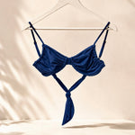 A navy blue strappy bikini top hanging on a white hanger from a gold clothing rack, set against a warm beige wall with soft leaf shadows.