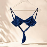 A navy blue strappy bikini top hanging on a white hanger from a gold clothing rack, set against a warm beige wall with soft leaf shadows.